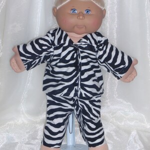 Pode incluir: Uma boneca Cabbage Patch Kid vestindo um pijama de zebra preto e branco. A boneca tem cabelo loiro, olhos azuis e uma tez rosada.