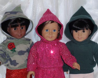 May include: Three dolls wearing hooded sweatshirts. The doll on the left is wearing a camouflage hooded sweatshirt with a red rose embroidered on the front. The doll in the middle is wearing a pink sequined hooded sweatshirt. The doll on the right is wearing a dark green hooded sweatshirt.