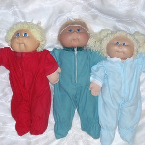 May include: Three Cabbage Patch Kids dolls wearing pajamas. The doll on the left is wearing red pajamas, the doll in the middle is wearing teal pajamas, and the doll on the right is wearing light blue pajamas.
