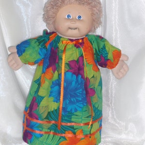 Pode incluir: Uma boneca Cabbage Patch Kid vestindo um vestido de estampa floral verde, laranja e azul com acabamento de fita laranja.