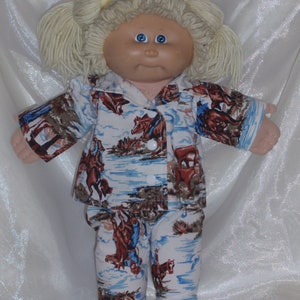 Pode incluir: Uma boneca Cabbage Patch Kid vestindo um pijama azul e branco com estampa de cowboy e cavalo. A boneca tem cabelo loiro e olhos azuis.