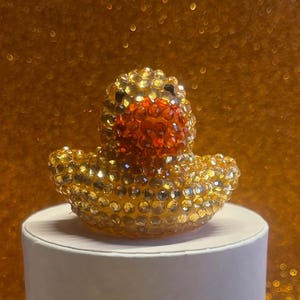 Puede incluir: Una figura decorativa de pato de goma enjoyada. El pato est&aacute; cubierto de pedrer&iacute;a dorada y transparente, con un pico naranja y ojos negros. Se encuentra sobre un pedestal blanco contra un fondo dorado brillante.