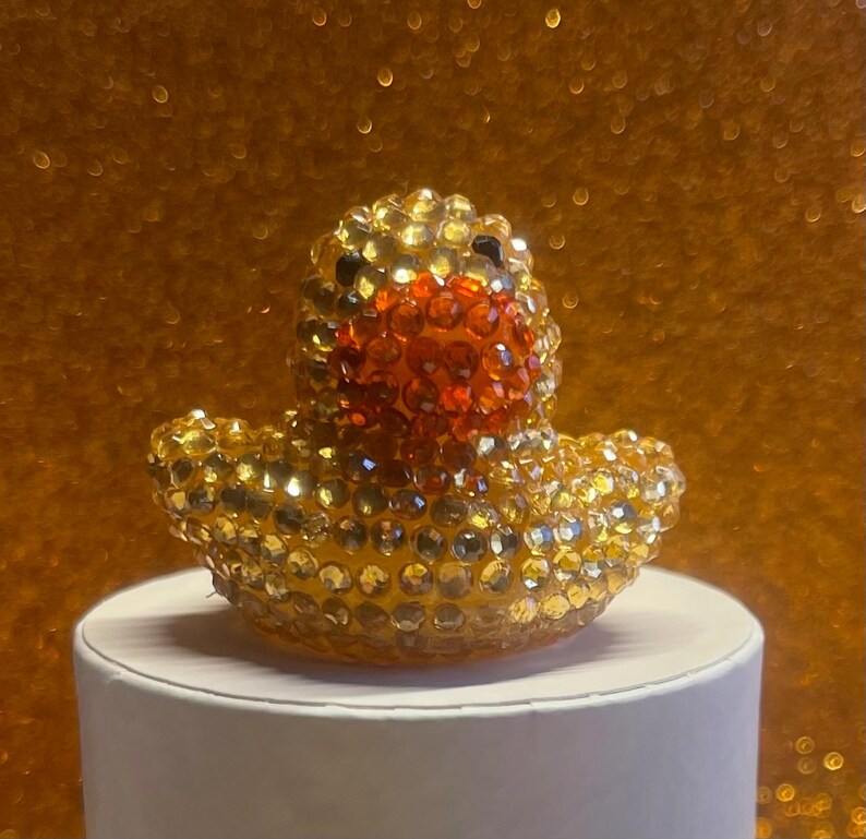 Puede incluir: Una figura decorativa de pato de goma enjoyada. El pato est&aacute; cubierto de pedrer&iacute;a dorada y transparente, con un pico naranja y ojos negros. Se encuentra sobre un pedestal blanco contra un fondo dorado brillante.