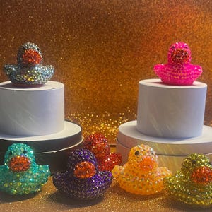 Puede incluir: Una colecci&oacute;n de figuras de patos de goma enjoyados en varios colores, incluyendo turquesa, morado, rojo, amarillo y plateado. Algunos patos se exhiben sobre pedestales blancos. El fondo es dorado y brillante.