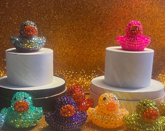 May include: A collection of bejeweled rubber duck figurines in various colors, including teal, purple, red, yellow, and silver. Some ducks are displayed on white pedestals. The background is a shimmering gold.