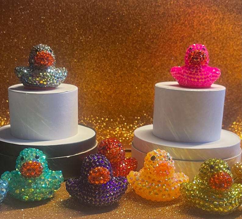 Puede incluir: Una colecci&oacute;n de figuras de patos de goma enjoyados en varios colores, incluyendo turquesa, morado, rojo, amarillo y plateado. Algunos patos se exhiben sobre pedestales blancos. El fondo es dorado y brillante.