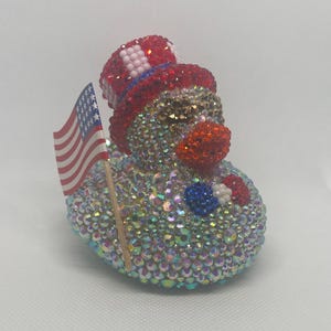 Puede incluir: Un pato de goma cubierto de cristales iridiscentes que lleva un sombrero de copa rojo, blanco y azul. El pato tiene un pico rojo, una pequeña pajarita azul y blanca y sostiene una bandera estadounidense en miniatura en un palo de madera.