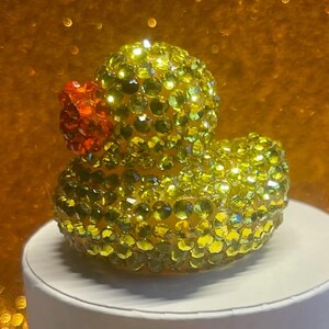 Puede incluir: Figura de pato de goma adornada con pedrer&iacute;a amarilla y verde, con pico naranja. La figura est&aacute; sobre una superficie blanca, con un fondo dorado borroso.