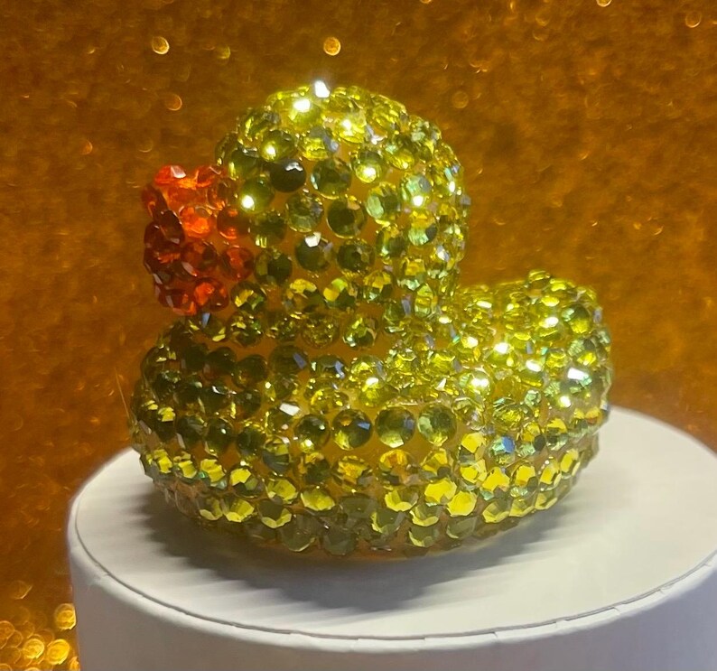 Puede incluir: Figura de pato de goma adornada con pedrer&iacute;a amarilla y verde, con pico naranja. La figura est&aacute; sobre una superficie blanca, con un fondo dorado borroso.