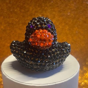 Puede incluir: Figura decorativa de pato cubierta de cristales negros y naranjas. El pato tiene ojos morados y un pico naranja. La figura est&aacute; sobre una superficie blanca con un fondo dorado.