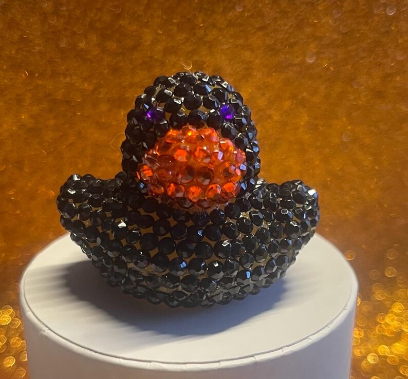 Puede incluir: Figura decorativa de pato cubierta de cristales negros y naranjas. El pato tiene ojos morados y un pico naranja. La figura est&aacute; sobre una superficie blanca con un fondo dorado.