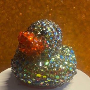 Puede incluir: Figura decorativa de pato cubierta de pedrer&iacute;a iridiscente multicolor. El pato tiene un pico naranja brillante y se encuentra sobre un fondo borroso marr&oacute;n dorado. Un art&iacute;culo decorativo &uacute;nico y llamativo.