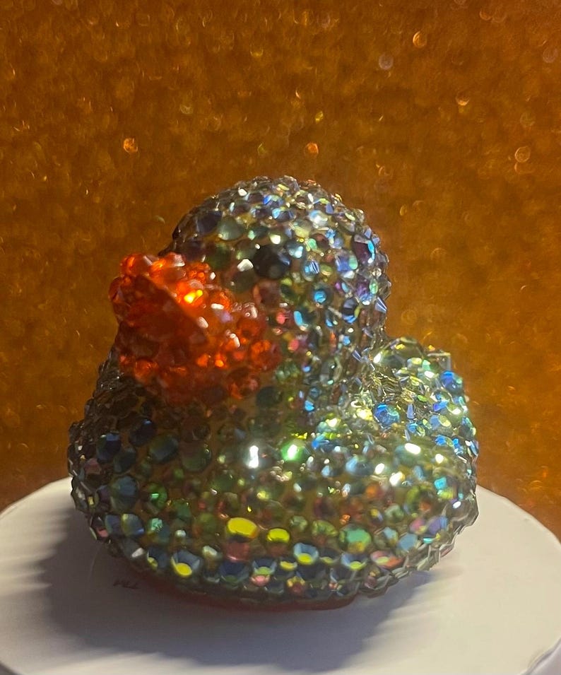 Puede incluir: Figura decorativa de pato cubierta de pedrer&iacute;a iridiscente multicolor. El pato tiene un pico naranja brillante y se encuentra sobre un fondo borroso marr&oacute;n dorado. Un art&iacute;culo decorativo &uacute;nico y llamativo.