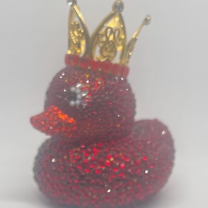 Puede incluir: Una figura de pato de goma completamente cubierta de pequeños cristales rojos brillantes. El pato presenta una corona de metal dorado con intrincados patrones recortados y pequeñas piedras transparentes en las puntas, sobre una banda de cristales rojos.