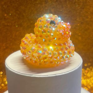 Puede incluir: Una figura decorativa de pato de goma enjoyado. El pato est&aacute; cubierto de cristales iridiscentes, naranjas y amarillos. Tiene una gema negra como ojo y est&aacute; sentado sobre una base cil&iacute;ndrica blanca. El fondo es de color dorado borroso.
