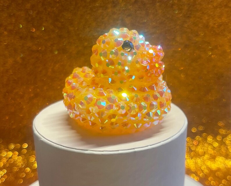 Puede incluir: Una figura decorativa de pato de goma enjoyado. El pato est&aacute; cubierto de cristales iridiscentes, naranjas y amarillos. Tiene una gema negra como ojo y est&aacute; sentado sobre una base cil&iacute;ndrica blanca. El fondo es de color dorado borroso.