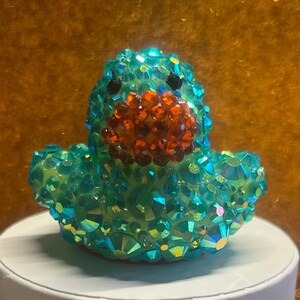 Puede incluir: Una figura de pato de color verde azulado adornada con pedrer&iacute;a naranja y negra. El pato est&aacute; cubierto de cristales iridiscentes, creando un efecto brillante. La figura es un objeto decorativo.