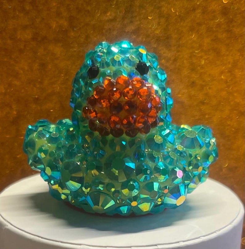 Puede incluir: Una figura de pato de color verde azulado adornada con pedrer&iacute;a naranja y negra. El pato est&aacute; cubierto de cristales iridiscentes, creando un efecto brillante. La figura es un objeto decorativo.
