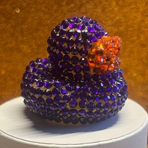 Puede incluir: Figura decorativa de pato cubierta de cristales facetados morados y naranjas. El pato tiene un cuerpo y una cabeza redondeados, con un pico naranja. Los cristales reflejan la luz, creando un efecto brillante. La figura se exhibe sobre una superficie blanca.