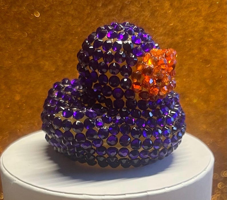 Puede incluir: Figura decorativa de pato cubierta de cristales facetados morados y naranjas. El pato tiene un cuerpo y una cabeza redondeados, con un pico naranja. Los cristales reflejan la luz, creando un efecto brillante. La figura se exhibe sobre una superficie blanca.