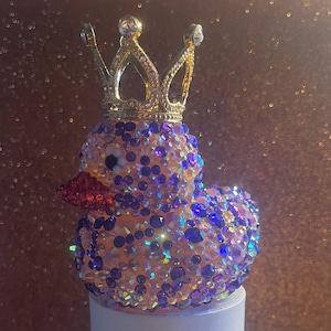 Puede incluir: Una figura de pato enjoyada con una corona dorada. El pato está cubierto de pedrería iridiscente en tonos rosa, morado y azul. El pico es rojo y los ojos son negros. La corona está adornada con cristales transparentes.