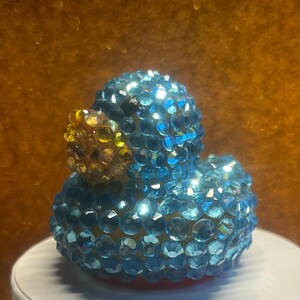 Puede incluir: Un pato de goma decorativo cubierto de brillantes cristales azules y transparentes. El pato tiene un pico amarillo y dorado. Est&aacute; sobre una superficie blanca, con un c&aacute;lido fondo dorado.