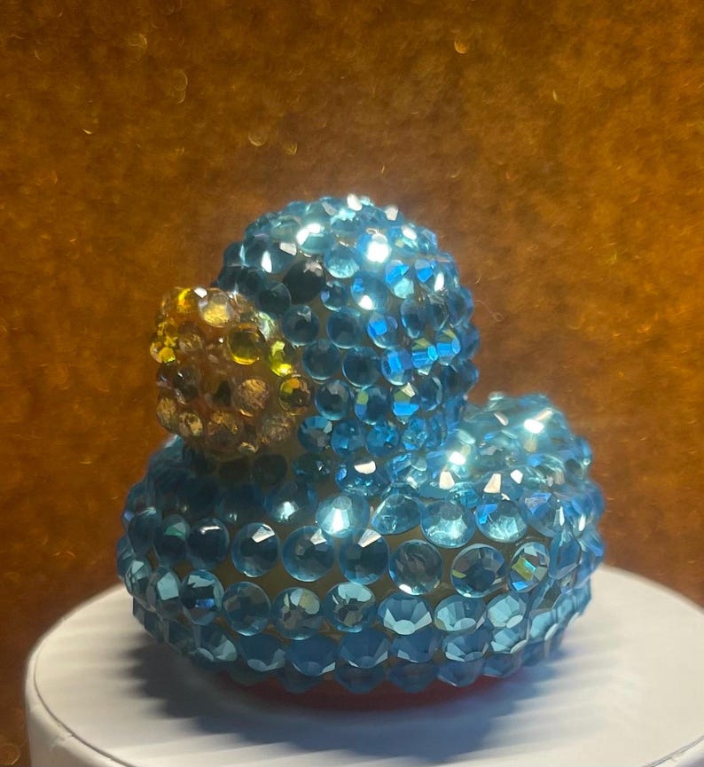 Puede incluir: Un pato de goma decorativo cubierto de brillantes cristales azules y transparentes. El pato tiene un pico amarillo y dorado. Est&aacute; sobre una superficie blanca, con un c&aacute;lido fondo dorado.