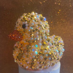 Puede incluir: Una figura de pato amarillo cubierta de cristales iridiscentes y dorados. El pato tiene un pequeño pico naranja y un ojo negro. La figura está sobre una superficie blanca contra un fondo dorado borroso.