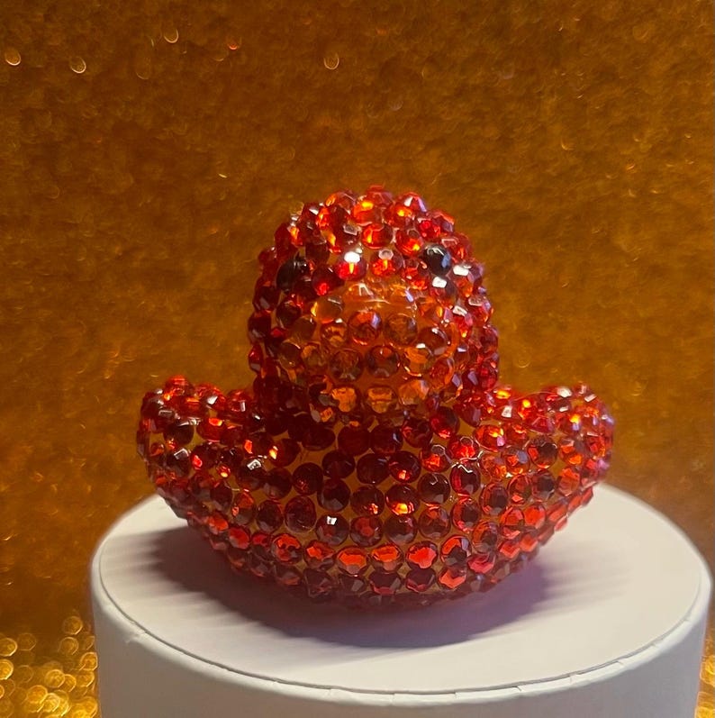 Puede incluir: Figura decorativa de pato cubierta de pedrer&iacute;a roja y naranja. El pato est&aacute; sobre una superficie blanca sobre un fondo dorado. La pedrer&iacute;a le da al pato un aspecto brillante.