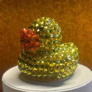 Puede incluir: Figura decorativa de pato de goma cubierta de pedrer&iacute;a amarilla y verde. El pato tiene un pico rojo y est&aacute; sobre una superficie blanca. El fondo es de color dorado c&aacute;lido.