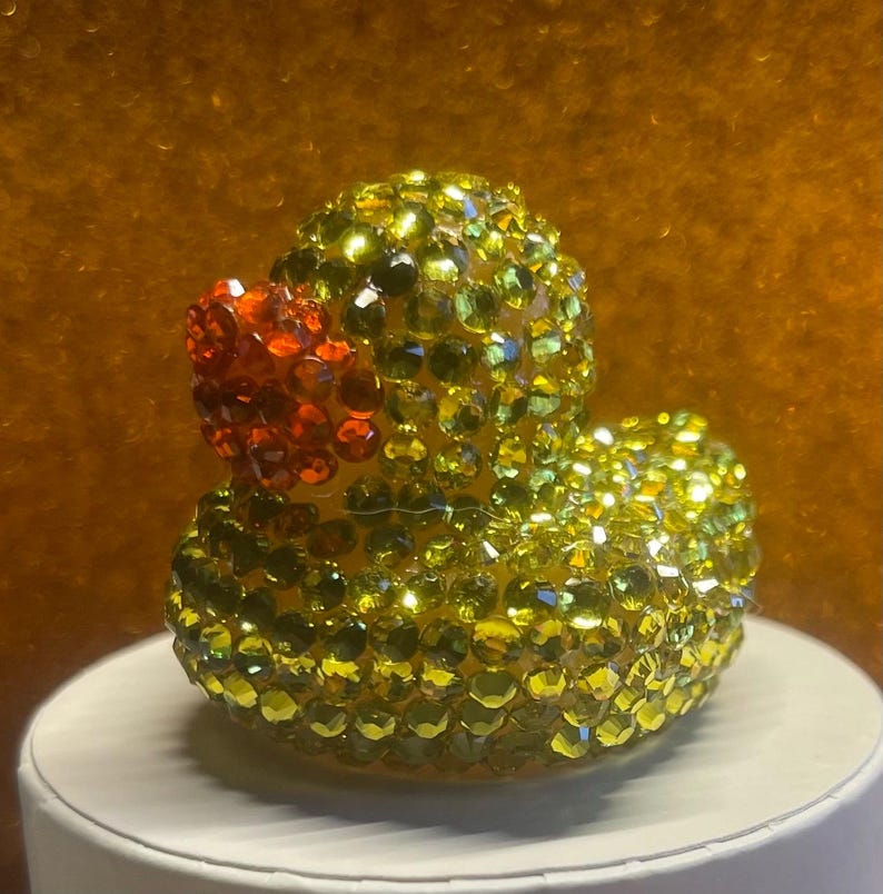 Puede incluir: Figura decorativa de pato de goma cubierta de pedrer&iacute;a amarilla y verde. El pato tiene un pico rojo y est&aacute; sobre una superficie blanca. El fondo es de color dorado c&aacute;lido.