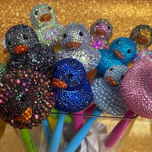 Puede incluir: Una colección de bolígrafos rematados con patitos de goma cubiertos de pedrería multicolor. Cada pato presenta un pico naranja brillante y ojos negros. Los bolígrafos tienen cuerpos de plástico de varios colores, incluyendo azul, rosa y verde.
