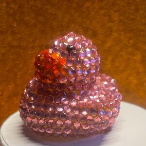 Puede incluir: Un pato de goma rosa enjoyado con un pico naranja brillante y un ojo negro. El pato est&aacute; cubierto de peque&ntilde;as gemas rosas brillantes. El fondo es de color naranja borroso.