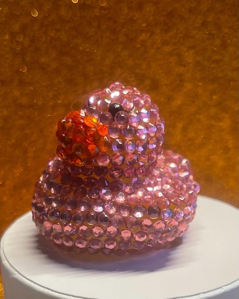 Puede incluir: Un pato de goma rosa enjoyado con un pico naranja brillante y un ojo negro. El pato est&aacute; cubierto de peque&ntilde;as gemas rosas brillantes. El fondo es de color naranja borroso.