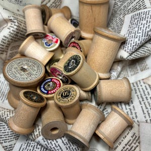 May include: A collection of vintage wooden spools, some with labels, resting on a fabric with printed text. The spools vary in size and have a natural wood color. Some labels read "Coats" and "Best Six Cord".