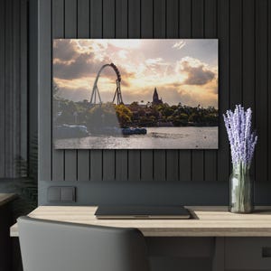 Könnte beinhalten: Gerahmter Druck einer Achterbahn vor einem Sonnenuntergangshimmel. Das Bild zeigt eine Loop-Achterbahn, einen See und Gebäude. Eine Vase mit Lavendel steht auf einem Schreibtisch vor dem Druck. Ein Laptop liegt auf dem Schreibtisch.