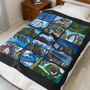 Puede incluir: Manta negra con un collage de imágenes de montañas rusas coloridas. La manta tiene un borde negro y se ven una almohada y una sábana blancas. La manta tiene el texto "The Howler" en azul y blanco.