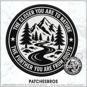 May include: A circular embroidered patch and a matching circular keychain featuring a black and white mountain landscape with pine trees and a winding road. The text reads: THE CLOSER YOU ARE TO NATURE THE FURTHER YOU ARE FROM IDIOTS.