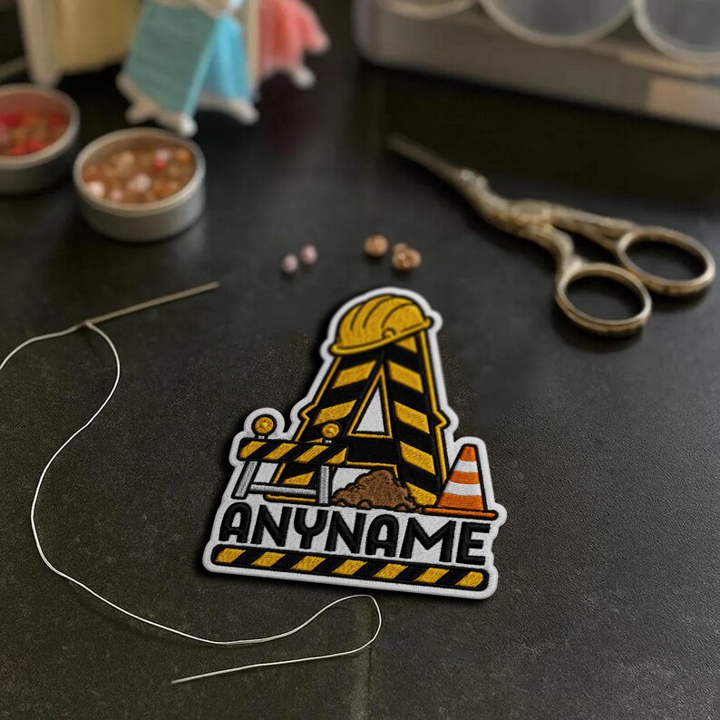 May include: An embroidered patch shaped like the letter A, designed with yellow and black hazard stripes, a yellow hard hat, a small pile of brown dirt, and an orange traffic cone. The text ANYNAME is embroidered in black at the bottom.