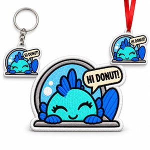 May include: A set featuring a keychain, an ornament with a red ribbon, and a patch, each displaying an embroidered turquoise fish in a gray bowl with a speech bubble containing the text HI DONUT! in black letters on a cream background.