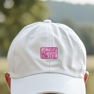Puede incluir: Gorra de b&eacute;isbol blanca con un parche rectangular rosa. El parche presenta las palabras "Spoiled WIFE" en una fuente decorativa, con coronas y corazones. La gorra tiene una visera curva y un bot&oacute;n en la parte superior.