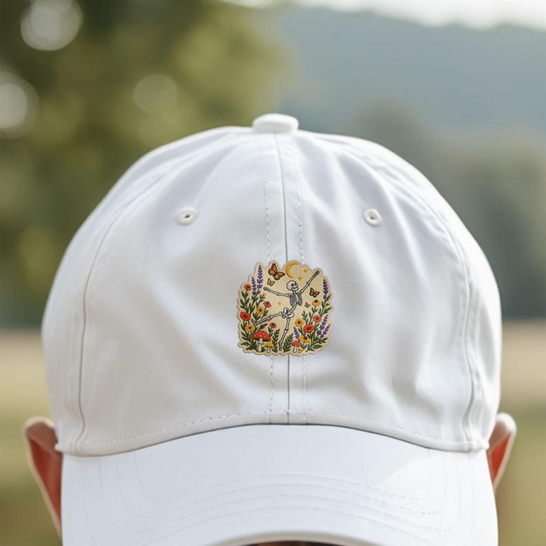 May include: A white baseball cap featuring a central embroidered patch. The patch depicts a skeleton dancing amidst colourful wildflowers, mushrooms, and butterflies under a crescent moon and stars, rendered in shades of orange, red, and purple.