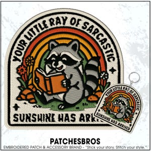 May include: Embroidered patch featuring a raccoon reading a book under a rainbow. The patch reads "YOUR LITTLE RAY OF SARCASTIC SUNSHINE HAS ARRIVED." A matching keychain is also visible. The patch is a fun accessory.