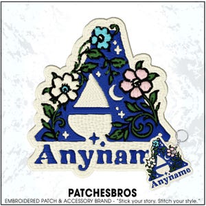 May include: An embroidered patch and matching keychain featuring the letter A in blue, decorated with green vines, white and pink flowers, a small crescent moon, and stars. The text Anyname is embroidered in blue below the letter A.