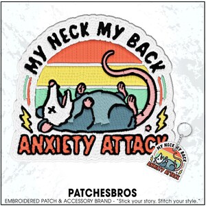 May include: Embroidered patch with a cartoon opossum design. The patch features the text "MY NECK MY BACK ANXIETY ATTACK" in a circular design with a sunset background. A matching keychain is also visible.