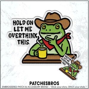 May include: Embroidered patch featuring a green frog wearing a cowboy hat and red bandana, holding a yellow mug. Text reads "HOLD ON LET ME OVERTHINK THIS." A smaller version of the design is attached as a keychain.