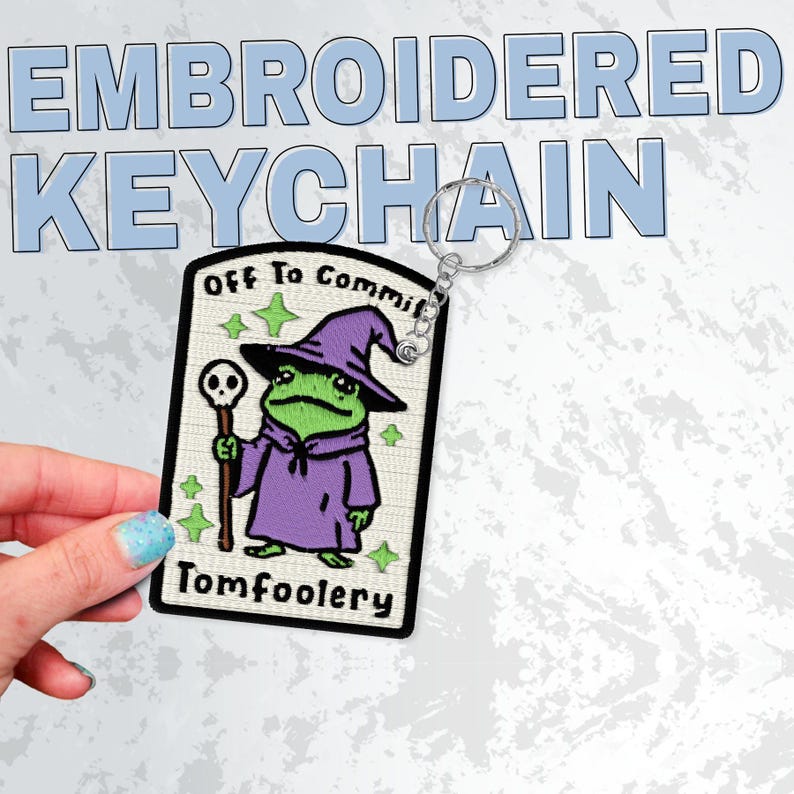 May include: Embroidered keychain featuring a cartoon frog wizard with the text "Off To Commit Tomfoolery." The frog wears a purple robe and hat, holding a staff with a skull. The keychain has a silver ring.