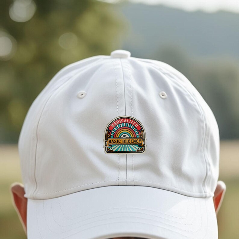 May include: A white baseball cap with a colorful patch that reads "RADICALIZED BY BASIC DECENCY." The patch features a rainbow design with red, orange, yellow, green, and blue colors. The cap is made of a smooth fabric and has a curved brim.