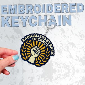May include: Embroidered keychain with a circular patch featuring a raised fist within a sunburst design, surrounded by a laurel wreath and text that reads "RADICALIZE DECENCY BY BASIC". The keychain has a silver metal ring.
