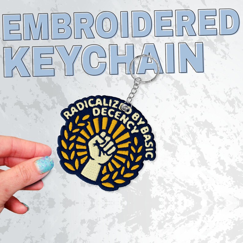May include: Embroidered keychain with a circular patch featuring a raised fist within a sunburst design, surrounded by a laurel wreath and text that reads "RADICALIZE DECENCY BY BASIC". The keychain has a silver metal ring.